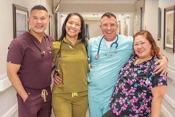 Four smiling medical professionals stand in a hospital hallway, wearing colorful scrubs and stethoscopes, conveying a sense of teamwork and positivity.