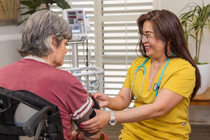 A nurse in a yellow uniform is smiling at an older patient in a wheelchair while taking their blood pressure. Medical equipment is visible in the background.