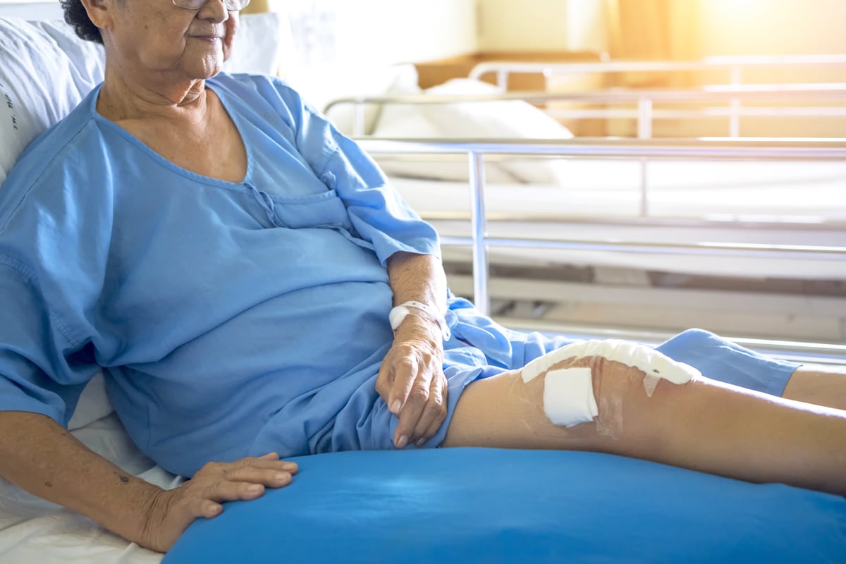 Elderly patient in a hospital bed with an IV, wearing a blue gown. The patient’s knee is bandaged, and warm sunlight streams through a nearby window.