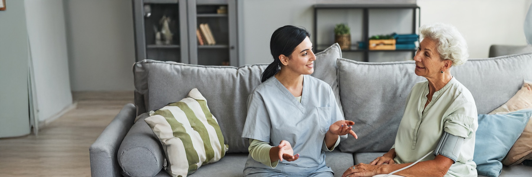 A caregiver in scrubs sits on a sofa, engaging warmly with an elderly woman in casual attire.